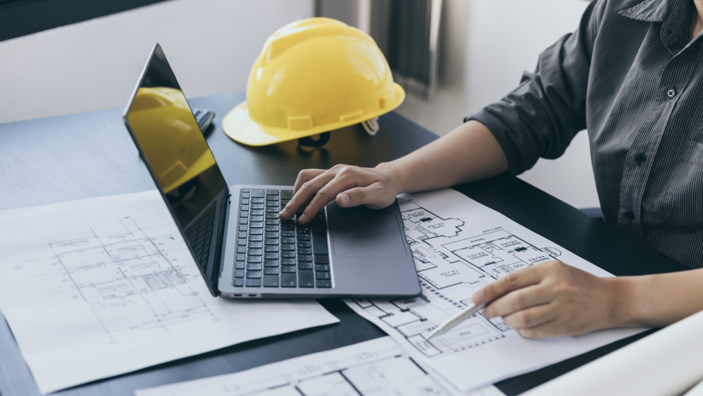 A person typing on a laptop while reviewing blueprints, with a yellow hard hat placed nearby.